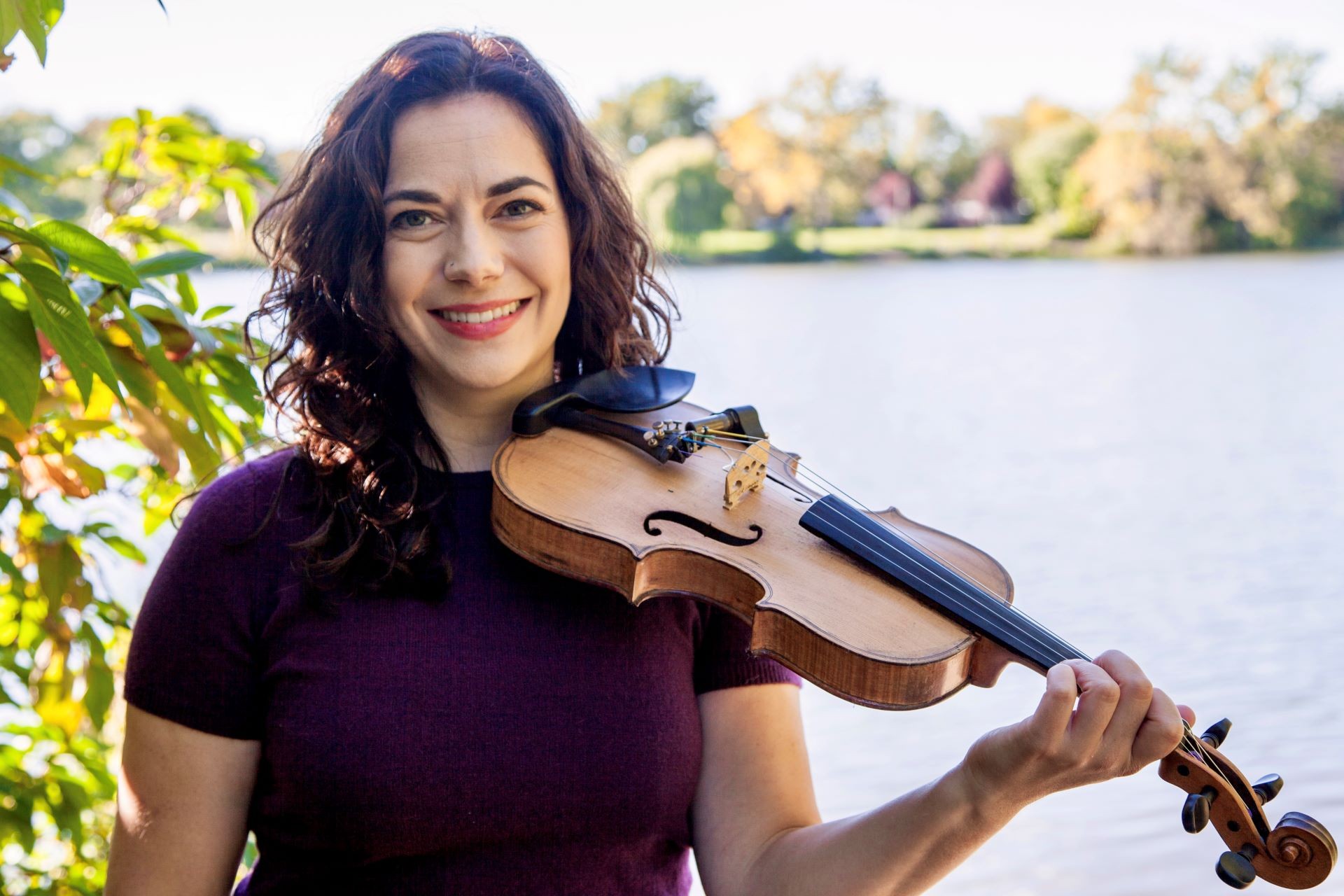 Une femme souriante, se tenant près d'un cours d'eau à l'extérieur, avec un violon. 