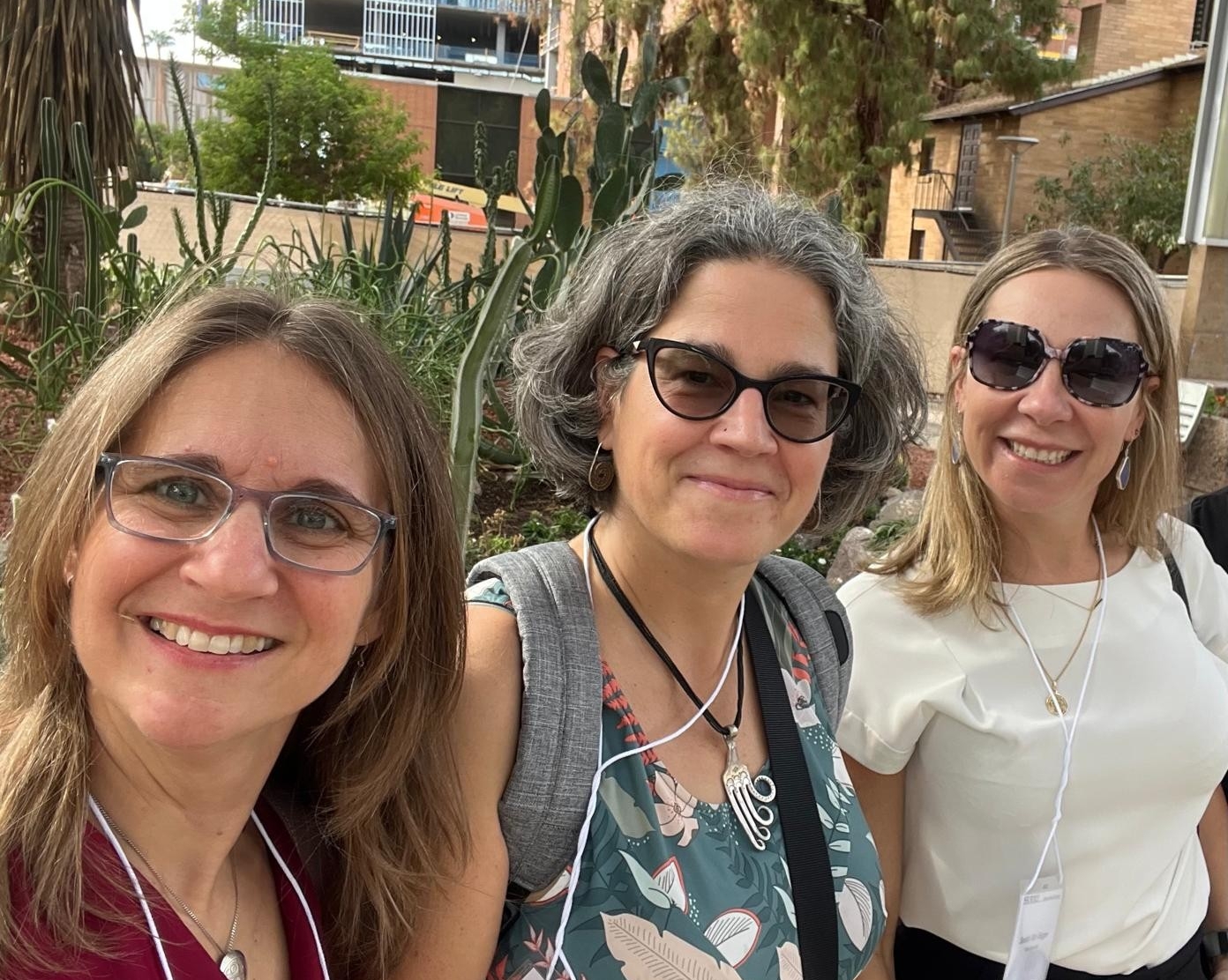 Trois femmes portant des lunettes de soleil, debout à l'extérieur, devant des cactus et diverses plantes.