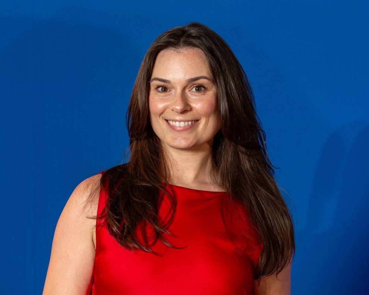 Une jeune femme souriante, aux cheveux longs et bruns, portant une robe rouge sur un fond bleu. 