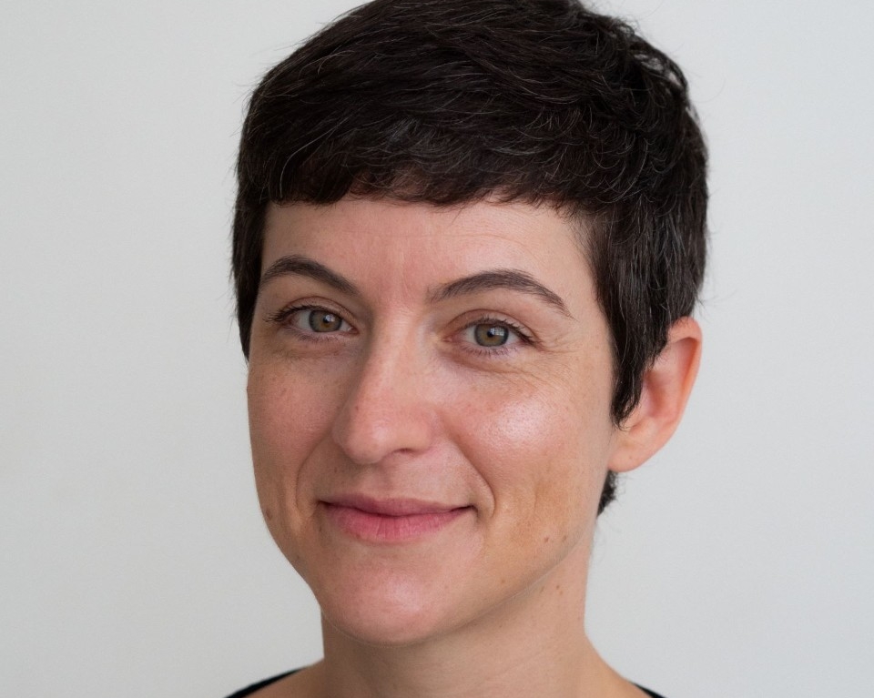 Une jeune femme souriante, aux cheveux bruns mi-longs, avec une frange, devant un arrière-plan flou où on peut aperçevoir des individus