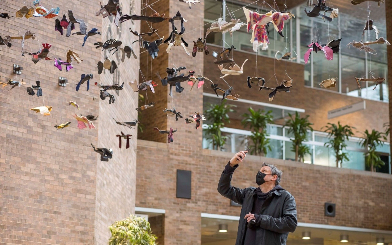Un homme portant un masque tient un appareil photo alors qu'il prend une photo d'oiseaux en papier suspendus avec un immeuble d'habitation en arrière-plan.