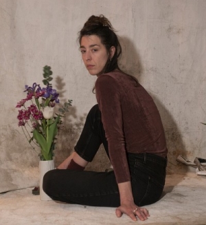 Portrait d'une femme aux longs cheveux noirs, portant un haut en velours brun et un pantalon noir, photographiée avec un vase de fleurs