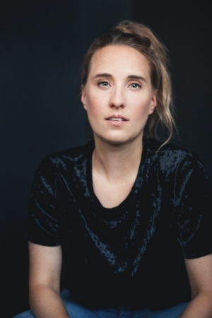 Photo d'une femme avec un t-shirt noir et des cheveux blonds en queue de cheval qui regarde l'appareil photo devant un mur sombre.