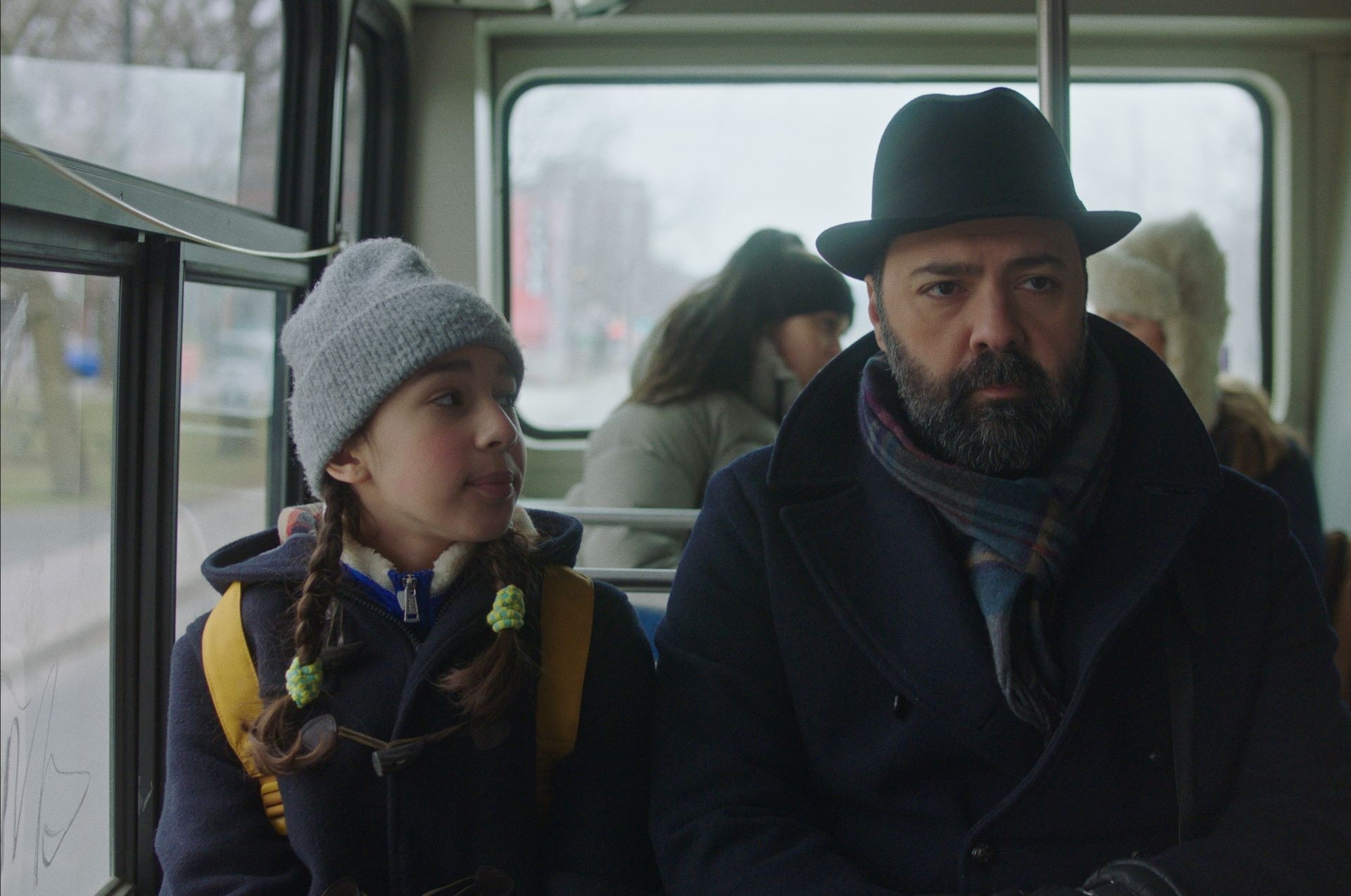 Une fille et un homme vêtus de vêtements d’hiver sont assis dans un bus. La jeune fille porte une tuque grise et regarde l'homme qui porte un chapeau noir.