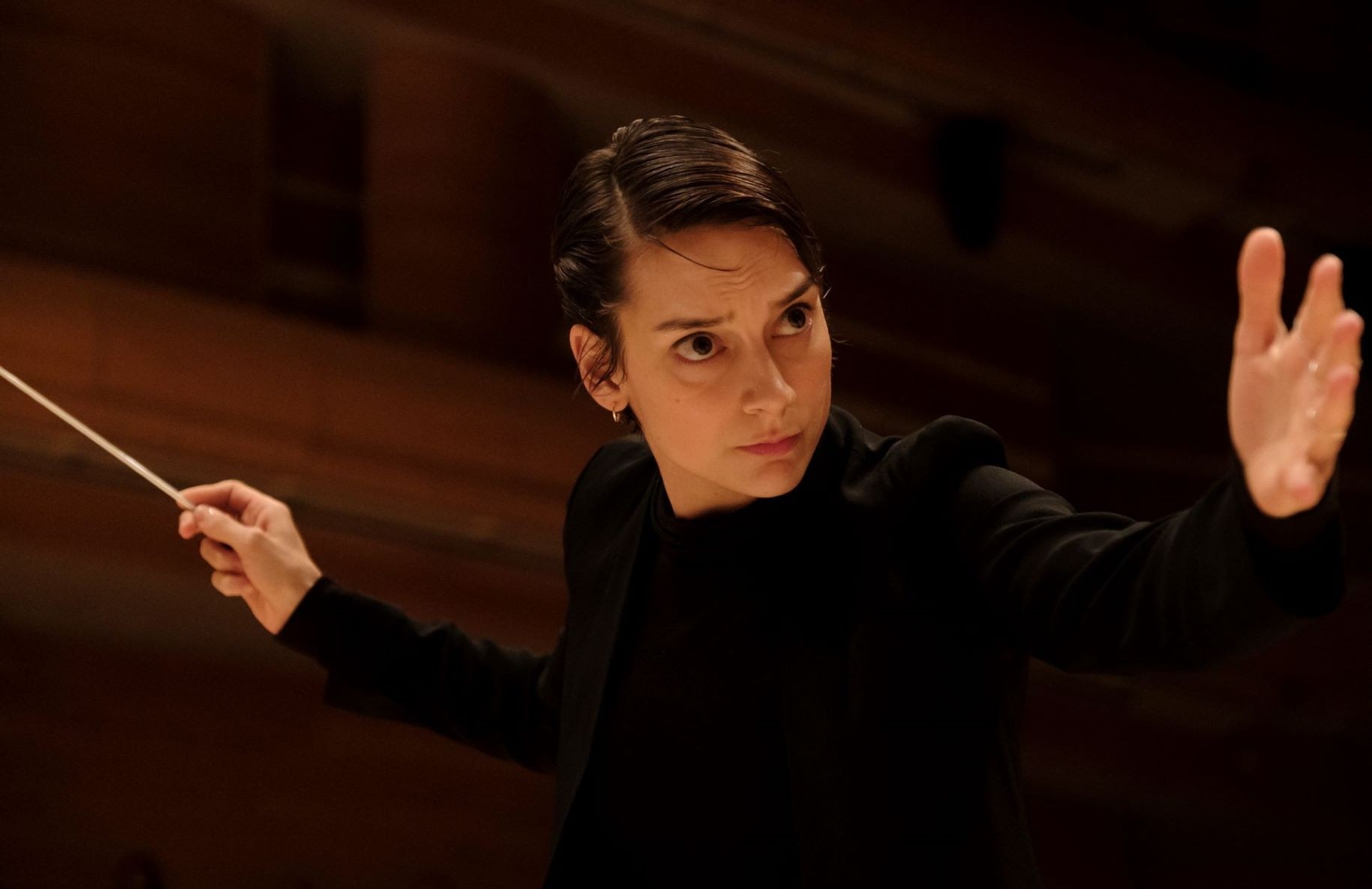 Cette image présente une femme concentrée aux cheveux courts et foncés, dirigeant un orchestre. Elle porte une tenue noire, les manches légèrement retroussées jusqu'aux avant-bras, et elle tient la baguette de chef d'orchestre dans sa main droite. Son regard intense et sa main gauche tendue suggèrent qu'elle est profondément engagée dans la direction de la performance, capturant un moment d'expression artistique et de leadership.