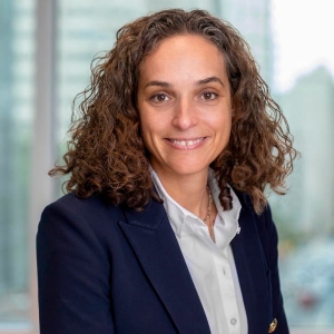 Portrait d'une femme souriante aux cheveux bouclés mi-longs portant un blazer bleu marine et un chemisier blanc à col.