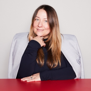 Une femme aux longs cheveux blonds-bruns porte un col roulé noir et un blazer gris clair drapé sur les épaules. Elle s'appuie sur une table rouge.