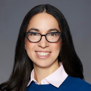 Portrait d'une femme souriante portant des lunettes à monture noire. Elle porte un pull bleu et un chemisier rose pâle.