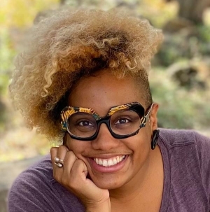 Portrait d'une femme souriante avec des lunettes à monture épaisse noire et orange et une chemise violette.