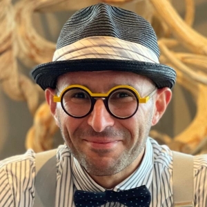 Un homme aux lunettes rondes noires et jaunes porte un fedora gris, une chemise rayée, des bretelles blanches et un nœud papillon noir.