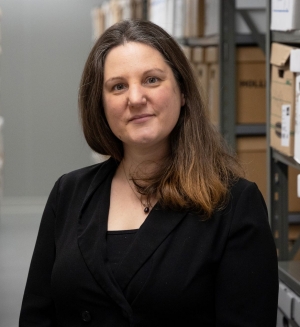 Une femme se tient dans une salle d'archives remplie de boîtes étiquetées, ce qui donne une impression d'organisation et de préservation.