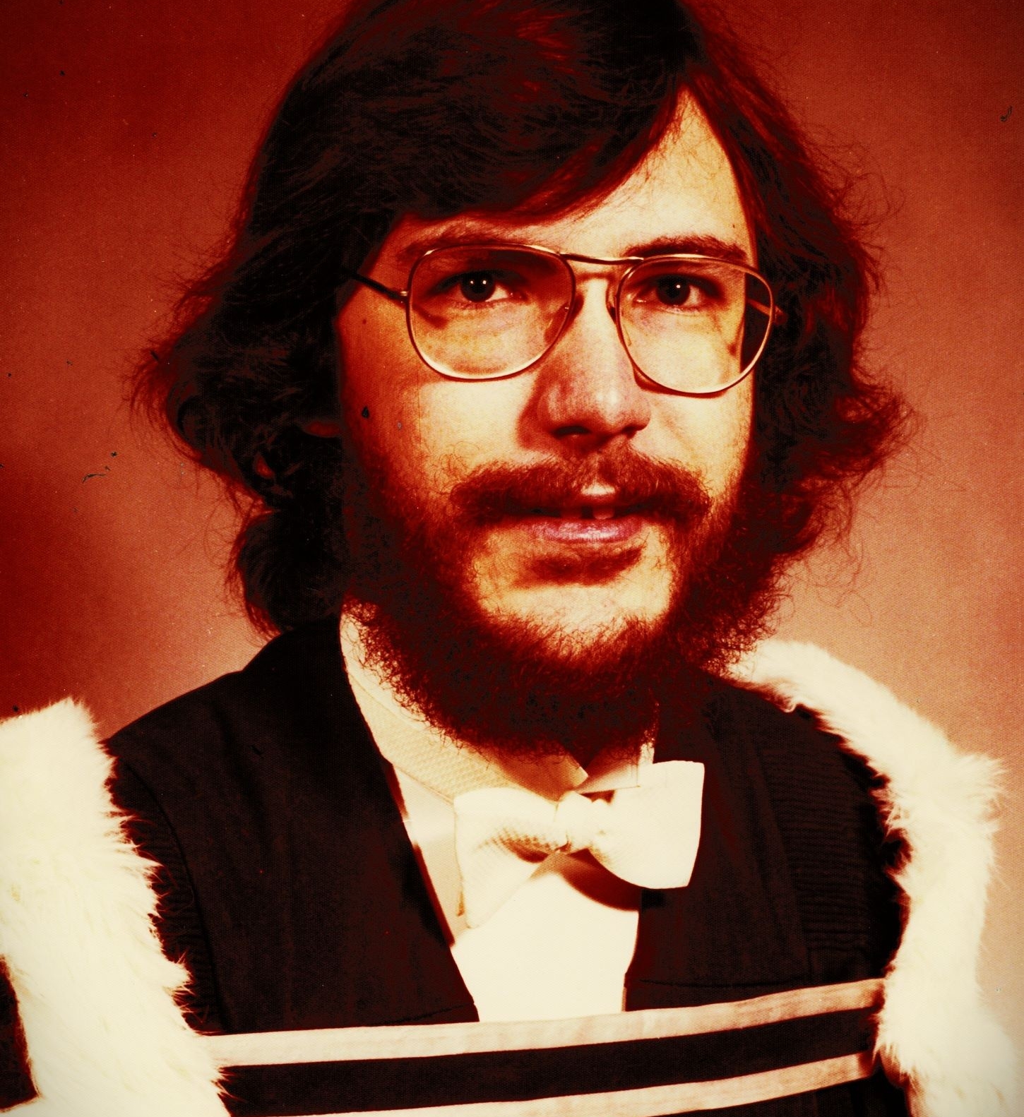 Portrait d'un homme aux longs cheveux bruns et à la barbe fournie, portant des lunettes et une robe de fin d'études avec un nœud papillon blanc. 