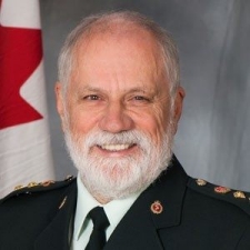Homme barbu aux cheveux courts, portant une chemise blanche avec une cravate et une veste foncées, sur fond de mur gris et d'une partie d'un drapeau canadien.