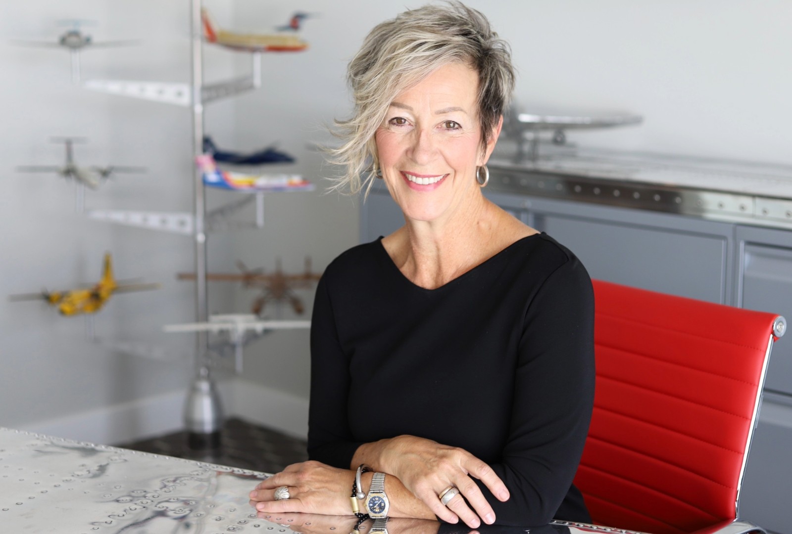 Tracy Medve Une femme aux cheveux courts gris-blancs est assise sur une chaise rouge dans un bureau. Elle a les bras croisés à son bureau et porte un pull noir à col en V.