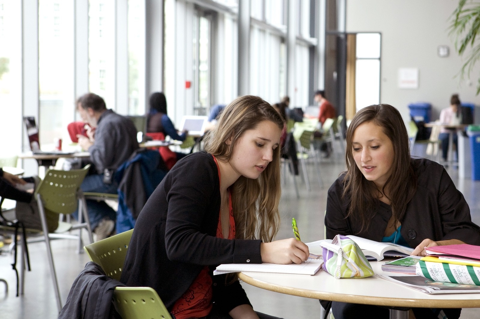Deux étudiantes étudient à une table dans un couloir lumineux avec d'autres étudiants assis à des tables à l'arrière-plan.