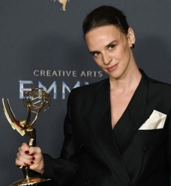 Une femme aux cheveux foncés tirés en arrière porte un blazer noir avec une pochette blanche et tient un Emmy Award.