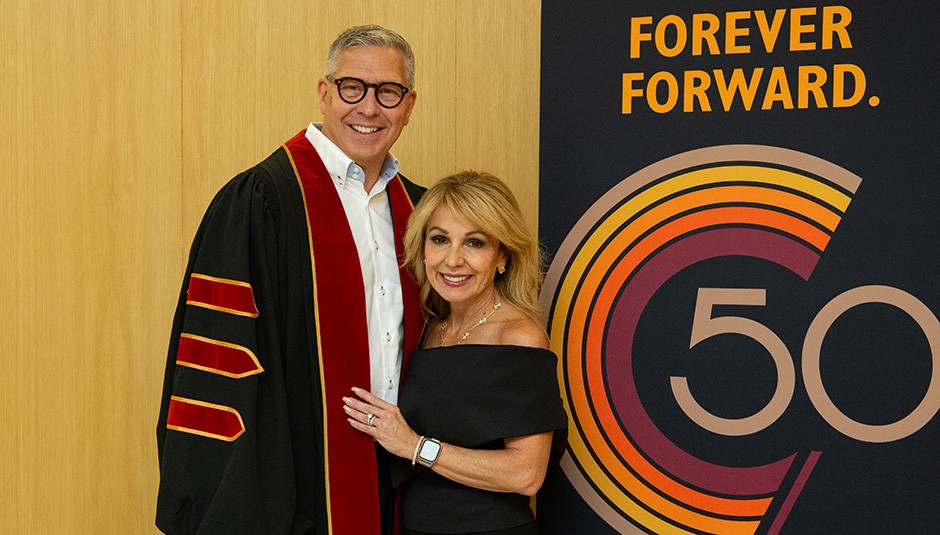 John porte une toge de diplômé et sa femme se tient fièrement à ses côtés.