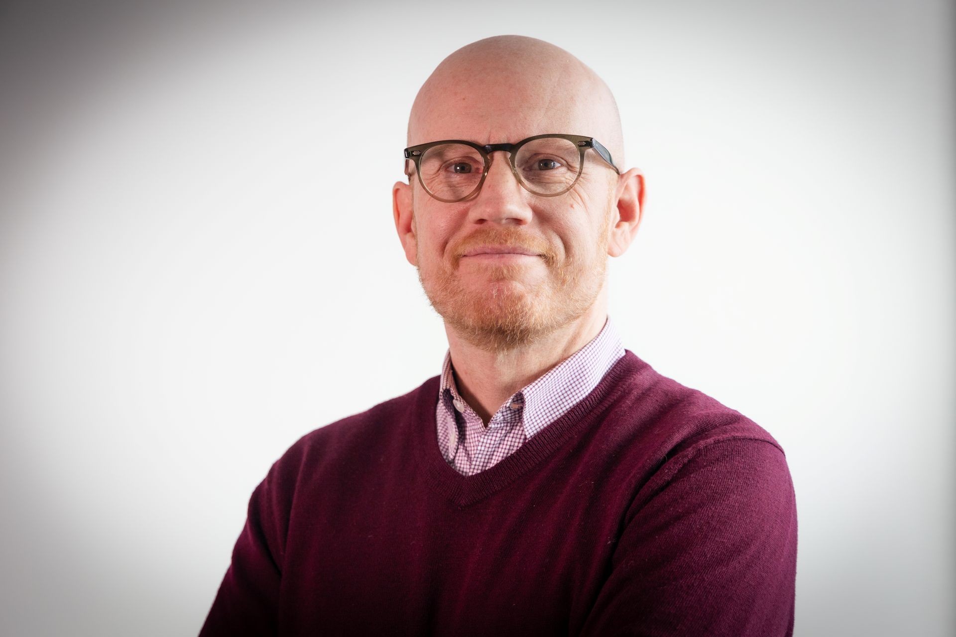 Un homme porte des lunettes et un pull bordeaux avec une chemise à carreaux rouges en dessous.