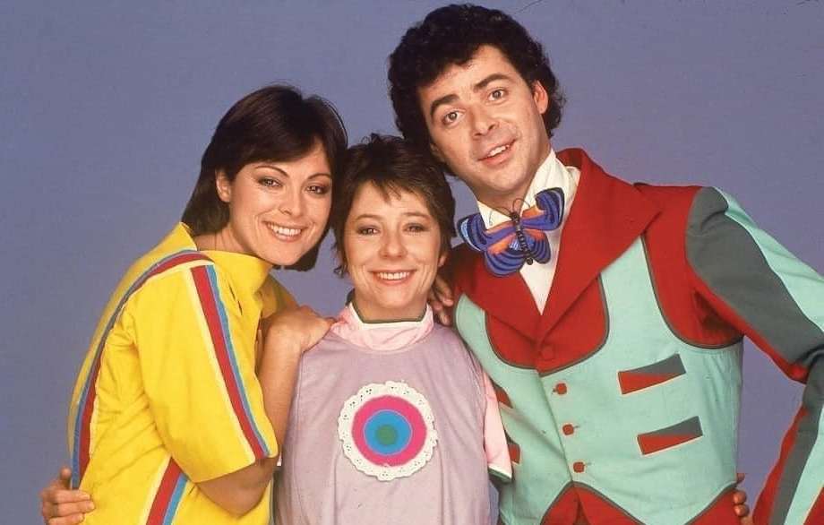 Trois adultes souriants posent côte à côte devant un fond bleu. La personne à gauche porte un haut jaune vif avec des rayures colorées sur les manches. La personne au milieu porte une tenue violet pastel avec un motif circulaire multicolore sur la poitrine. La personne à droite porte un costume coloré avec un gilet couleur menthe, des accents rouges et une cravate en forme de papillon. Tous trois ont les bras autour les uns des autres. 