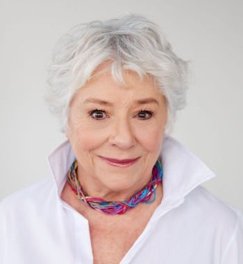 Femme aux cheveux blancs, vêtue d'un chemisier blanc et d'un collier coloré, sur fond blanc.