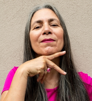 Skawennati fait le « signe de la paix » avec sa main droite sous son menton. Elle porte un t-shirt rose et a les cheveux longs.