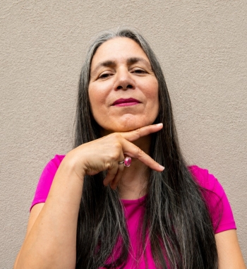 Skawennati fait un « signe de paix » avec sa main droite sous son menton. Elle porte un t-shirt rose et a les cheveux longs.