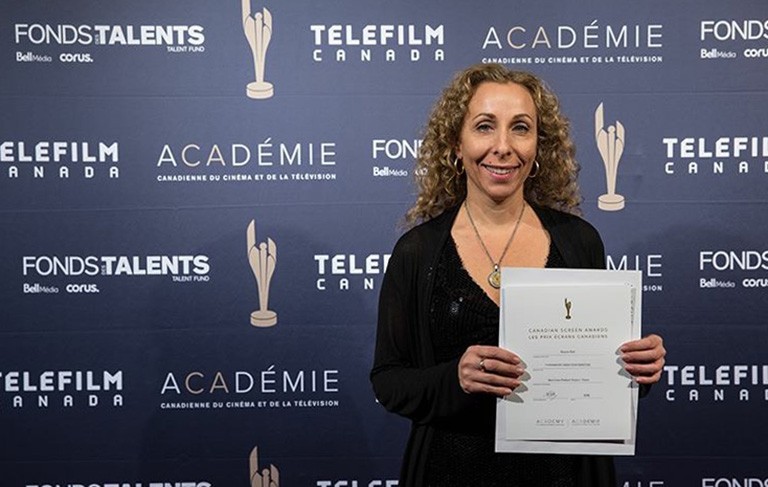 Rosana se tient devant une toile de fond sur laquelle figure le logo de Téléfilm Canada et brandit sa fiche de mise en candidature.