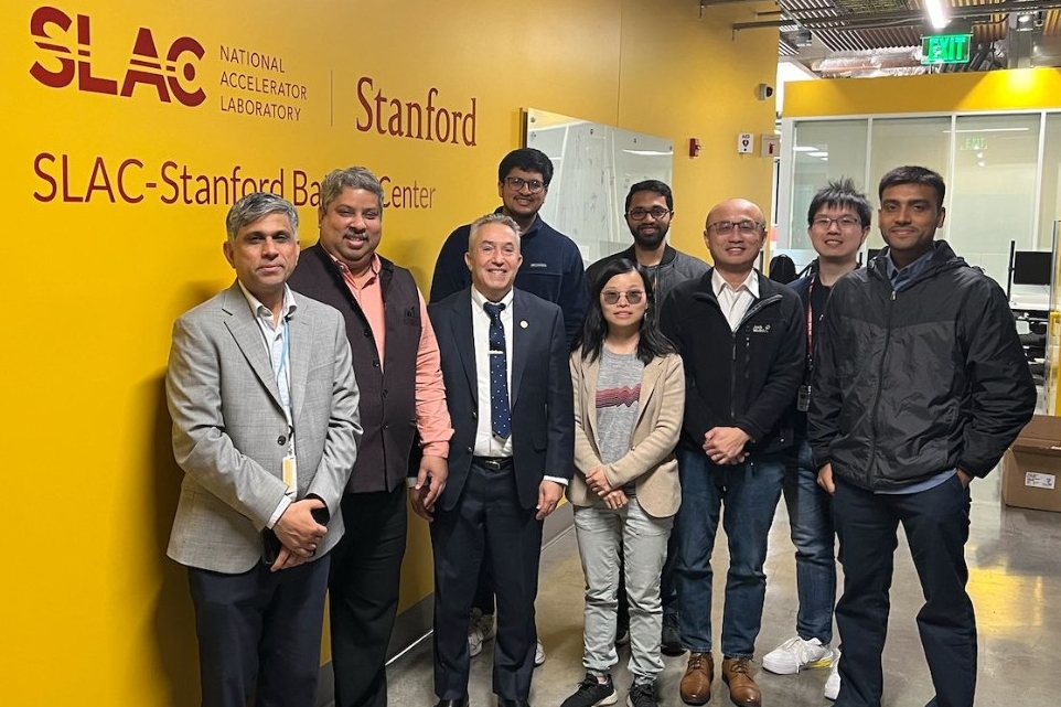 Un groupe de scientifiques pose devant un mur jaune portant les mots "SLAC National Accelerator Laboratory Stanford" et, en dessous, les mots "SLAC-Stanford Battery Center".