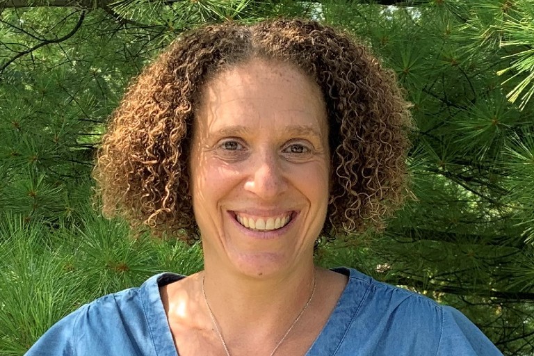 Elana Bloom : « En plus de motiver le personnel et d’encourager la mobilisation, la présence physique favorise la santé mentale et le bien-être. » Une femme aux cheveux bouclés et mi-longs sourit et se tient devant de la verdure.