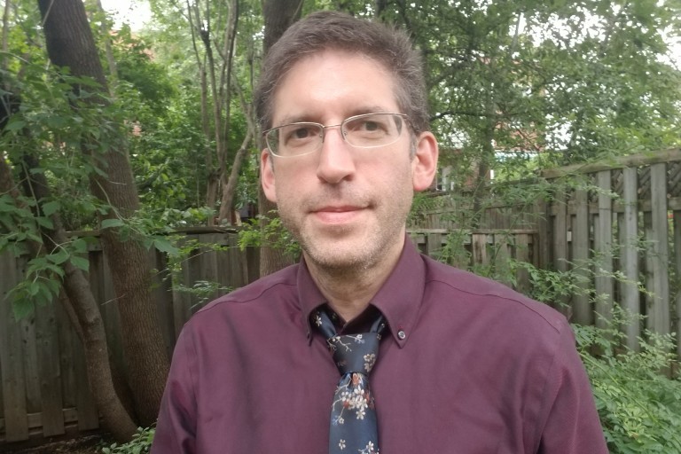 Charles Altman : « Nous avons fait la transition vers les activités en ligne et nous ferons la transition vers un mode hybride. » Un homme avec une cravate et des lunettes se tient devant une clôture, avec un peu de verdure en arrière-plan.