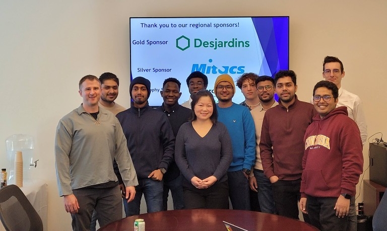 A group of students pose smiling in front of a projected image of sponsors. 