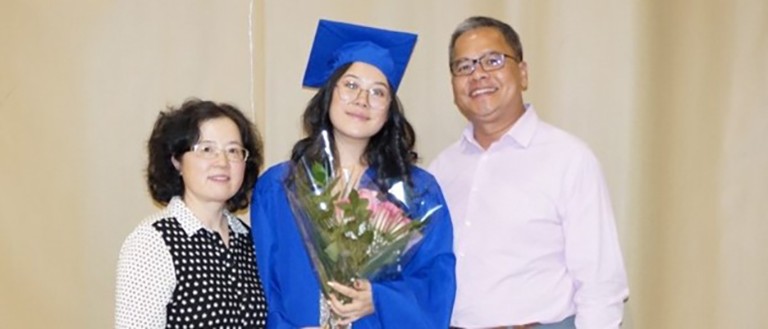 Une jeune femme portant sa tenue de fin d'études et se tenant aux côtés de ses parents.