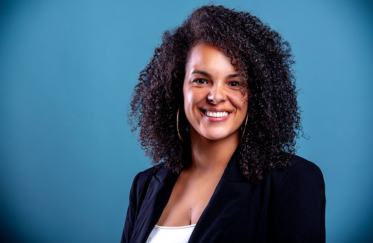 Jeune femme souriante aux longs cheveux noirs bouclés, vêtue d'un haut blanc et d'un blazer noir