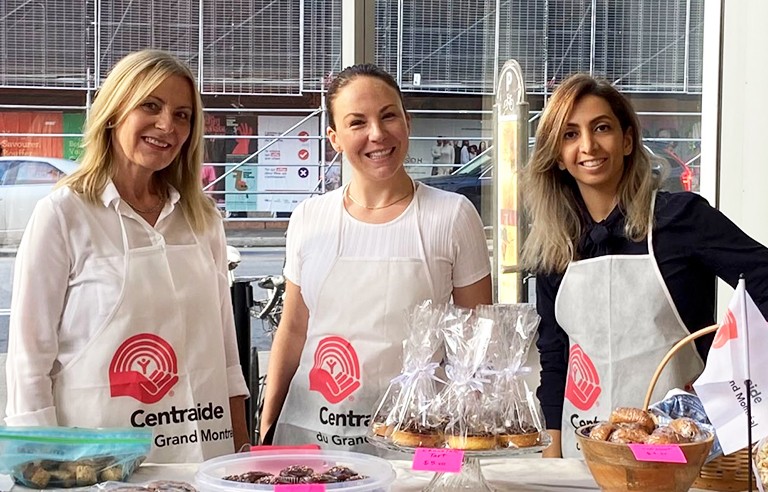 Trois femmes debout dans un espace intérieur, derrière une table remplie de produits de boulangerie, et portant des tabliers sur lesquels on peut lire "Centraide".