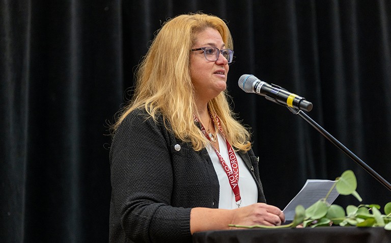 Une femme aux longs cheveux roux, portant des lunettes et un haut noir, s'exprime sur un podium.