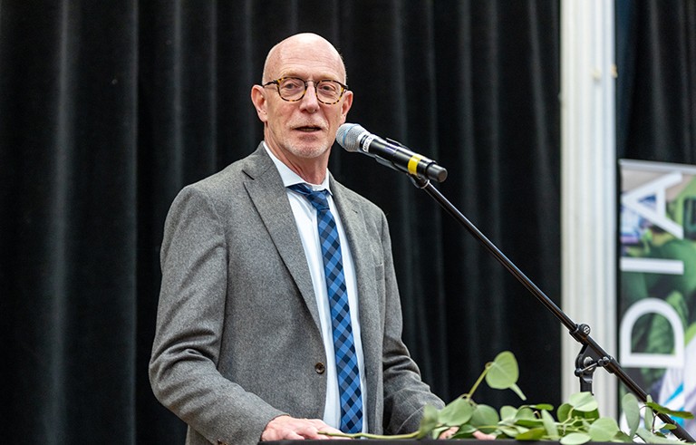 Un homme d'un certain âge, aux cheveux gris et courts, portant des lunettes, vêtu d'un costume et s'exprimant sur un podium.