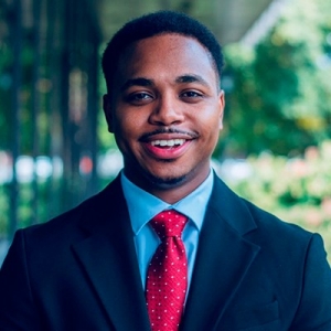 Jeune homme souriant aux cheveux noirs et courts, portant une barbe, une chemise bleue, une cravate rouge et un blazer bleu foncé.