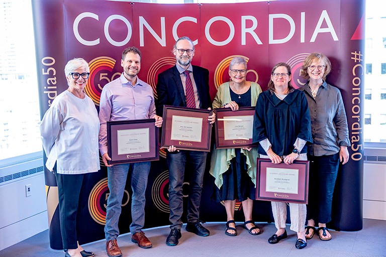 De gauche à droite : Anne Whitelaw, vice-rectrice exécutive aux affaires académiques, Noah Drew, Alex De Visscher, Lisa Ostiguy, Elizabeth Bloodgood et Kristina Huneault, vice-rectrice exécutive adjointe au développement du corps professoral et à l’inclusion. Un groupe de personnes, dont plusieurs tiennent des plaques, se tient à l'intérieur et sourit à l'appareil photo.