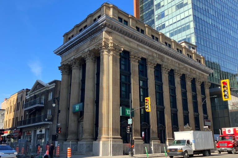Bâtiment historique avec de hautes colonnes à l'angle d'une rue de la ville, avec l'enseigne de la Banque TD. Une structure moderne en verre est visible à l'arrière-plan. Feux de circulation et véhicules présents.