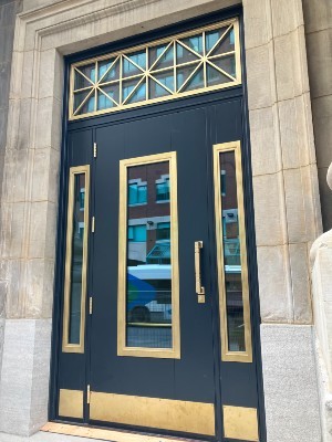 Une porte bleu foncé avec des accents en laiton et des détails géométriques sur l'imposte vitrée, située dans une façade de bâtiment en pierre.