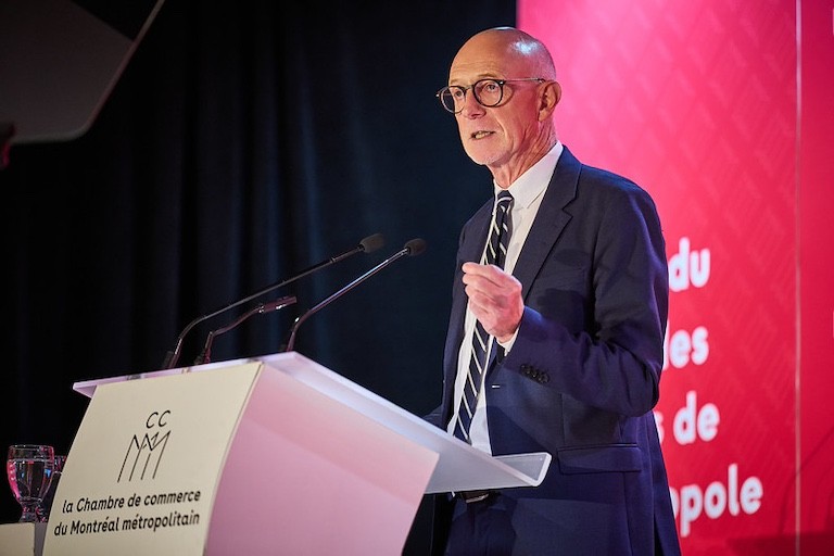 Un homme portant des lunettes et un costume d'affaires prononçant un discours derrière un podium blanc, devant un mur rouge et noir
