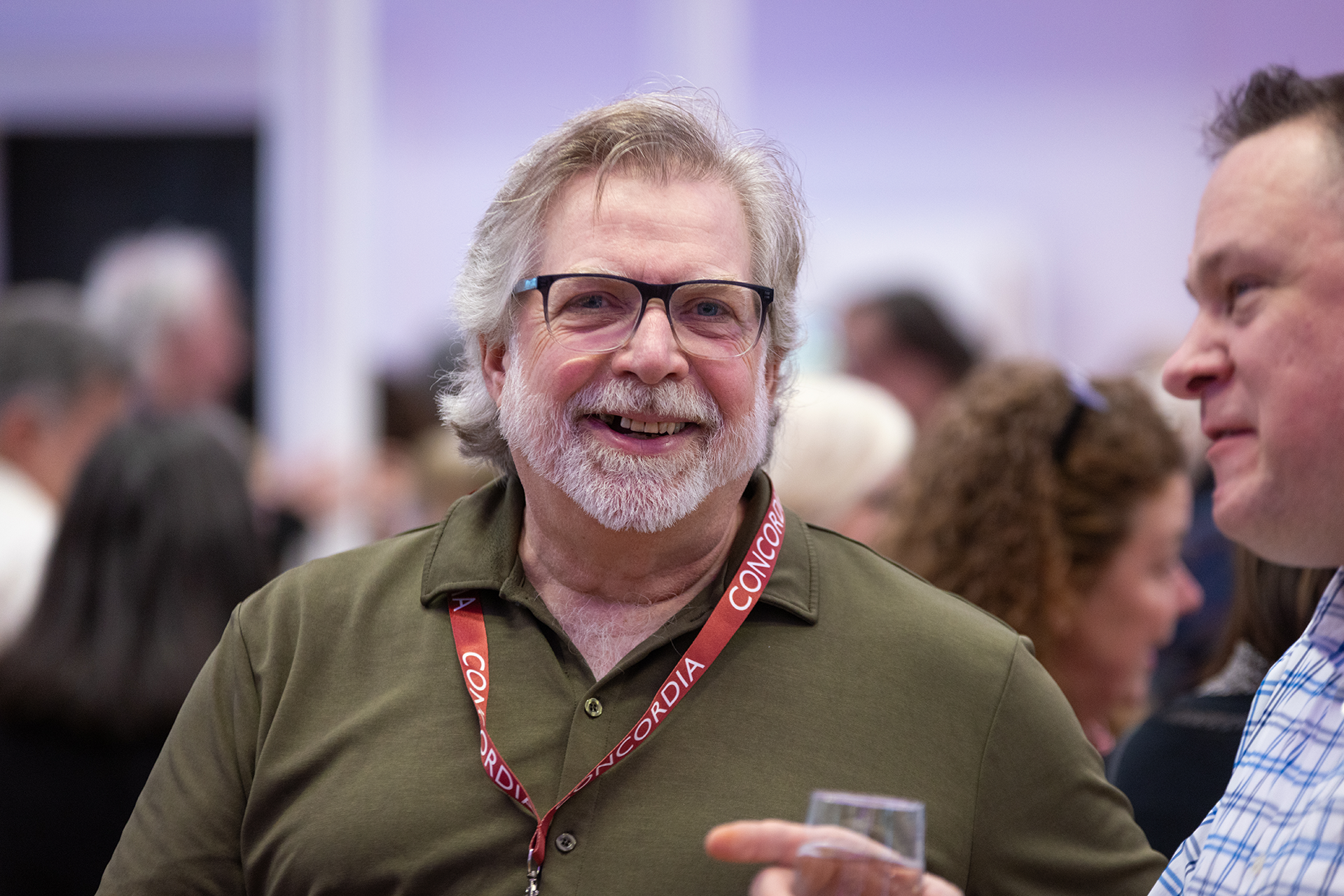 Un homme à la barbe grise et aux lunettes, avec un cordon autour du cou, sourit et discute avec un autre homme, partiellement visible sur la photo.