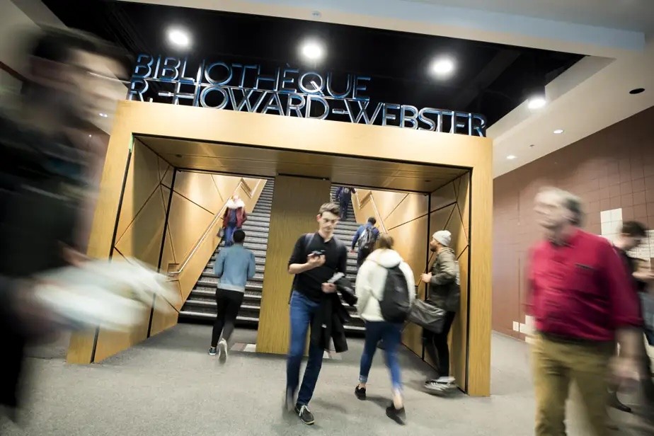 Des étudiants circulent à l’entrée de la bibliothèque, avec un escalier central et un passage encadré de bois moderne, tandis que des silhouettes floues au premier plan évoquent l’animation du campus.