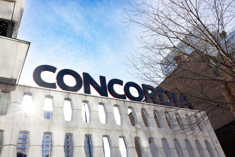 Grand panneau de Concordia sur la façade d’un pavillon du campus Sir-George-Williams; des bâtiments environnants et des branches d’arbres sont visibles sur fond de ciel bleu.