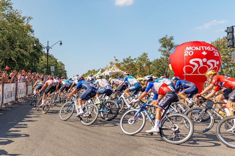 Des cyclistes roulent sur une rue de Montréal sous les yeux de spectateurs derrière des barrières, avec une affiche de l’événement cycliste Montréal 2026 en arrière-plan.