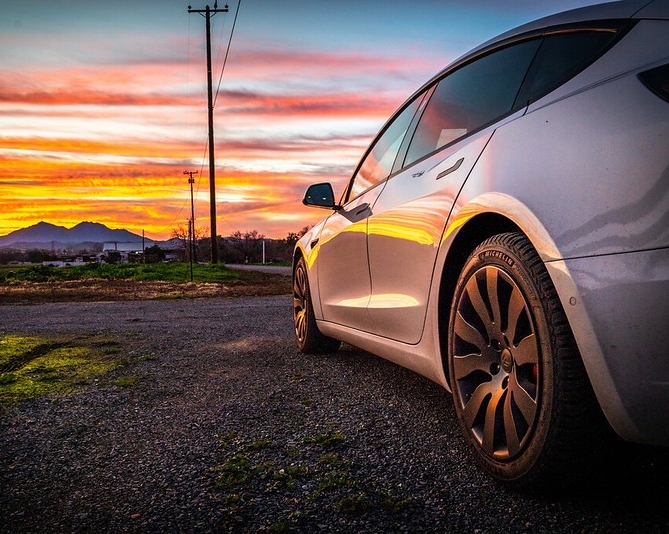 Voiture électrique stationnée sur une route de gravier face à un coucher de soleil et des montagnes lointaines